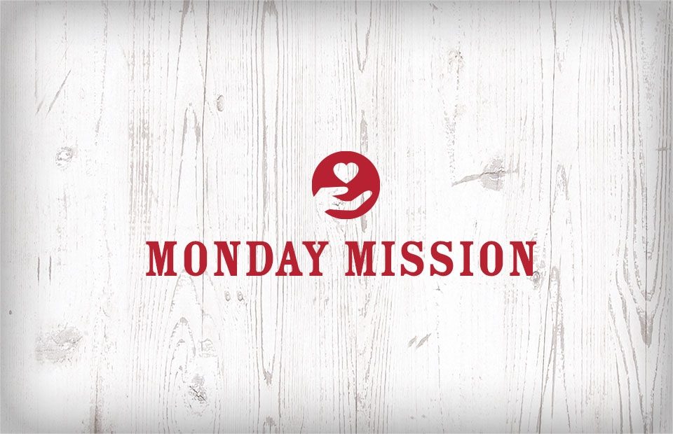 A white wooden background with a red circular logo featuring a hand holding a heart, and bold red text that reads "Monday Mission".