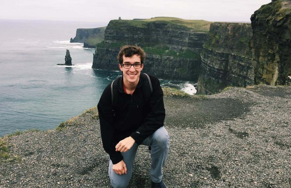 A young man with glasses, dark hair, and a black jacket crouches on a rocky cliff overlooking the ocean and green cliffs in the background.