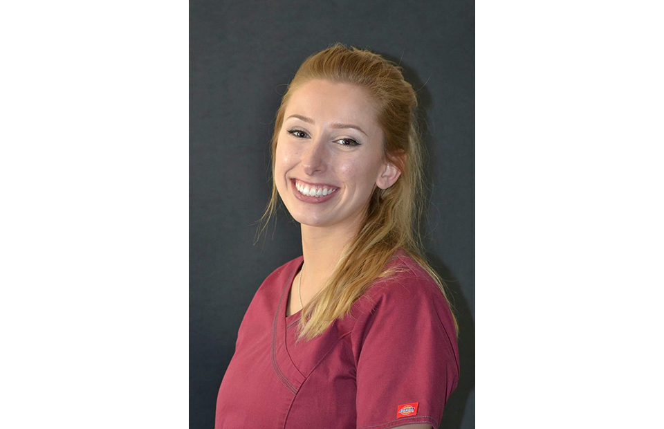 A smiling woman with long blonde hair wearing a maroon uniform with a Pizza Ranch patch on the sleeve, standing against a dark background.