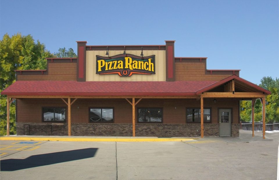 A single-story Pizza Ranch restaurant with a brick and wood exterior, large sign above the entrance, parking lot in front, and a clear blue sky.