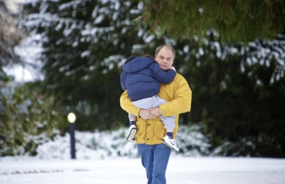A man in a yellow jacket carries a young child in blue clothing on his shoulders in a snowy outdoor setting with large evergreen trees in the background.
