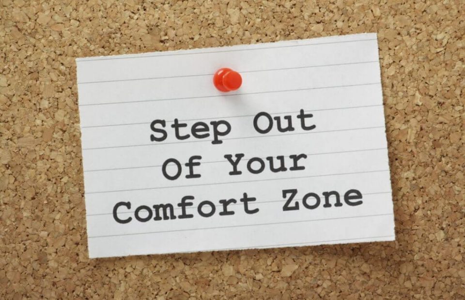 A note pinned on a corkboard with a red pushpin reads: "Step Out Of Your Comfort Zone" in black, bold letters on lined white paper.