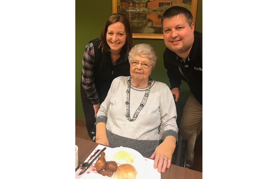 An elderly woman with white hair and glasses sitting at a table with a plate of food, flanked by a woman and a man standing beside her, all smiling.