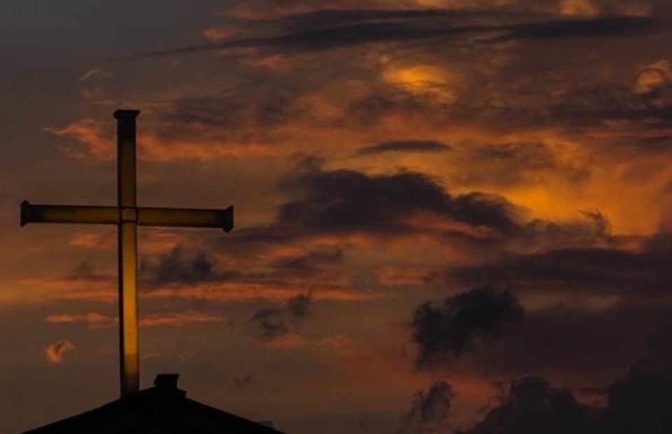 A cross mounted on a roof silhouette against a dramatic orange and dark clouded sunset sky.