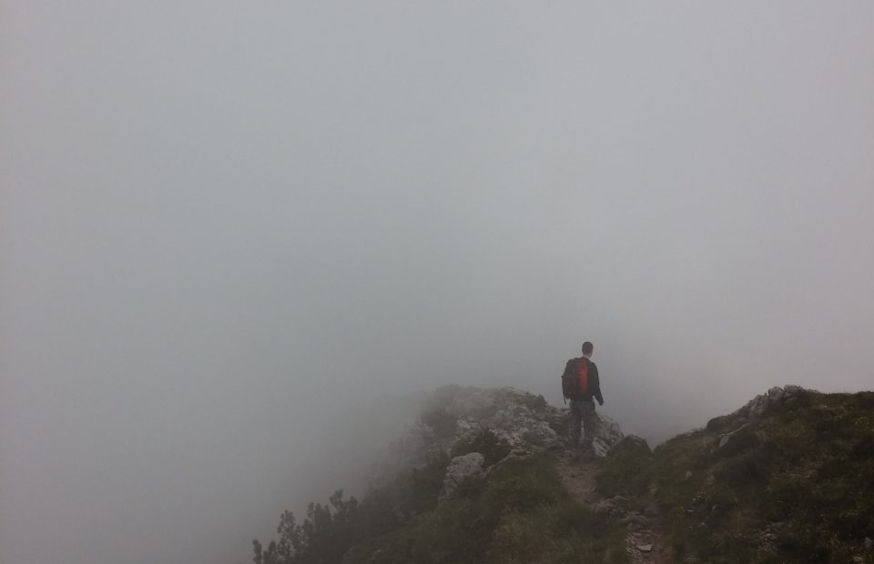 A person with a red backpack walking along a narrow mountain trail surrounded by fog and mist, with rocky terrain and sparse vegetation.
