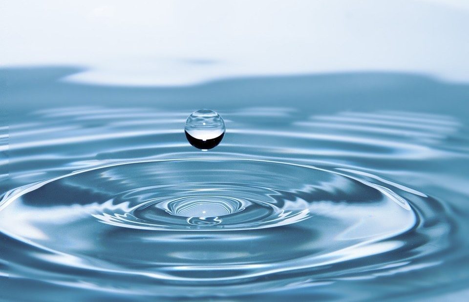 A single water droplet is captured mid-air just above a calm water surface, creating concentric ripples as it prepares to land.