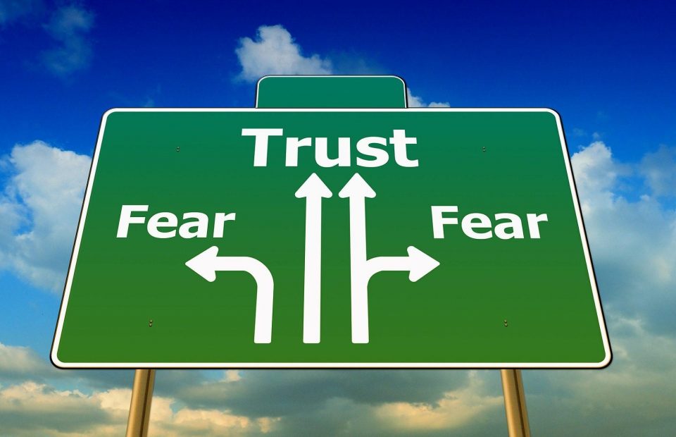 A green road sign with arrows pointing left, straight, and right labeled "Fear," "Trust," and "Fear," respectively, against a blue sky with clouds.