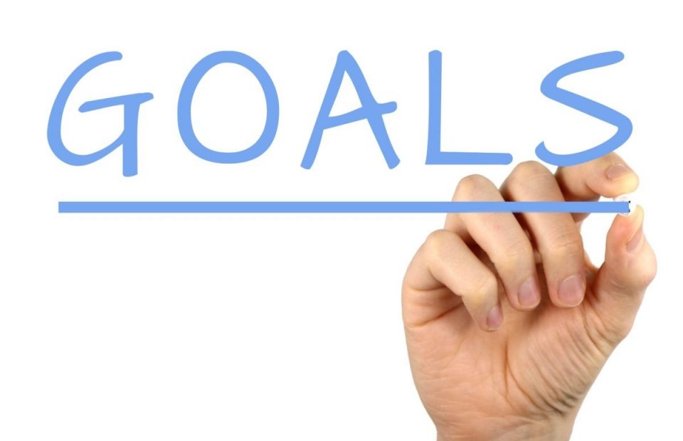 Handwriting the word "GOALS" in large blue letters on a transparent surface, with a hand holding a blue marker underneath.