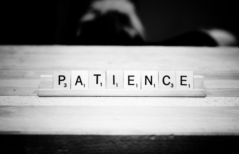 Scrabble tiles spelling "PATIENCE" on a wooden surface, with a blurred figure in a dark uniform in the background.