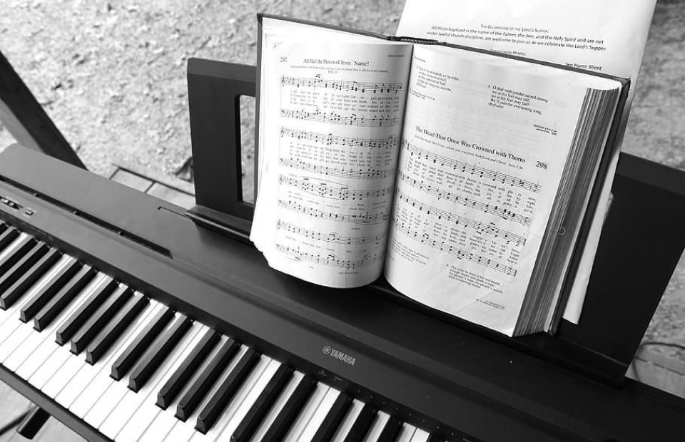 Open hymnal on a stand above a Yamaha keyboard, displaying sheet music with text and musical notes, outdoors on a paved surface.
