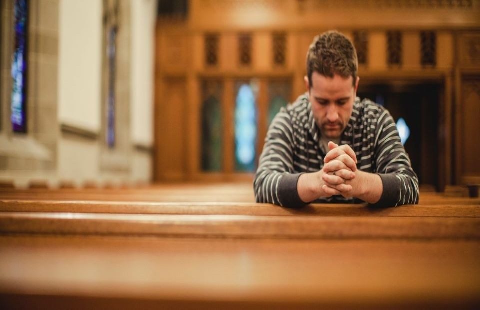 A man kneeling with his hands clasped in prayer on a church pew, with stained glass windows and wooden paneling in the background.