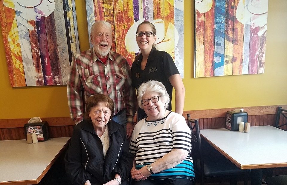 Four people, three women and one man, are inside a restaurant near a yellow wall with colorful abstract artwork behind them.